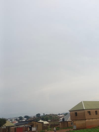 The image depicts a rural or semi-rural landscape, likely in the city of Rafin Bauna, Nigeria, under an overcast sky. In the foreground, a light brown, two-story building with a green corrugated metal roof and visible windows is prominent. To the left of this building, another structure with a dark roof is partially visible, along with a red metal gate or fence. In the middle ground and background, a collection of houses with various roof types, including some with corrugated metal, are spread across rolling hills dotted with trees. A single person is visible on a motorcycle in the lower right quadrant of the image, moving away from the viewer. The overall impression is one of a developing community with a mix of traditional and modern housing.