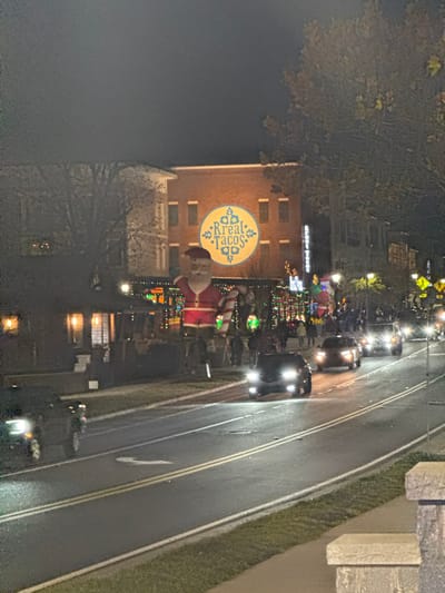 Night shot of a town street lined with Christmas lights. A building displays a circular "Real Tacos" sign and a large Santa Claus decoration. Cars drive on the right side of the road. Trees are partially visible on the left. The sky is dark, indicating nighttime. The overall scene conveys a festive holiday atmosphere.