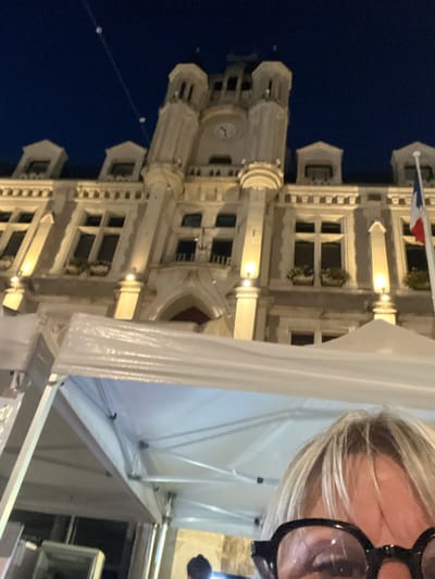 A low-angle, nighttime shot captures the facade of a historic building, likely the Hôtel de Ville in Saint-Galmier, France, illuminated by warm lights. The building features ornate architectural details, multiple windows, and a prominent clock tower with a visible clock face. A French flag flies from a pole on the right side of the building. In the foreground, a portion of a white tent structure is visible, partially obscuring the lower part of the building. A woman's face, wearing large black-rimmed glasses, is in the bottom right corner, looking upwards. The sky above is a deep, dark blue, with some small, illuminated elements that could be festive lights strung across the scene.