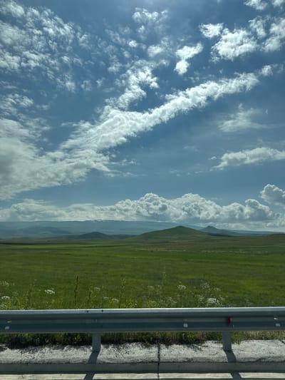 The image shows a scenic landscape with rolling green hills under a bright blue sky filled with fluffy white clouds. In the foreground, a silver guardrail runs horizontally across the frame, separating the viewer from a grassy verge dotted with wildflowers. In the midground, gently sloping hills create a sense of depth, and in the distance, mountains are visible, some with patches of snow. The overall impression is one of a vast, open, and natural environment. The city of Lanjik, Armenia, is known for its picturesque mountainous terrain and lush green valleys, which aligns with the scenery depicted in the image.
