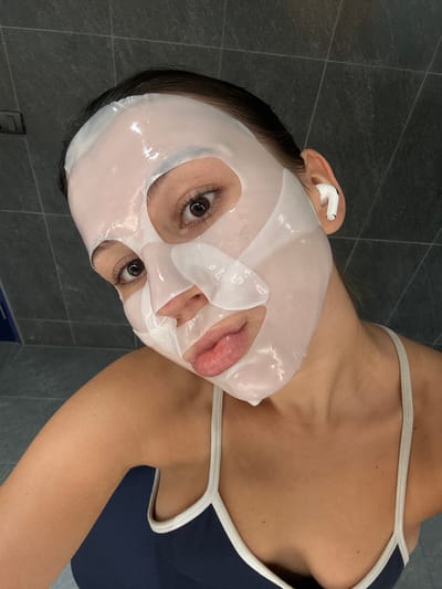A young woman with dark hair is wearing a white sheet face mask and white wireless earbuds. She is looking at the camera with a slight smile and her eyes are open. The woman is wearing a dark blue tank top with white trim. The background appears to be a bathroom with dark gray tiled walls. The image is a close-up selfie. The location is not provided.
