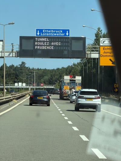 The image depicts a view from inside a vehicle traveling on a highway in Niederanven, Luxembourg. Overhead electronic signs display traffic information. The first sign, in blue, reads "Ettelbruck Lorentzweiler" with a downward arrow. Below this, a larger digital display shows "TUNNEL" in the top line and "ROULEZ AVEC PRUDENCE" in the second line, indicating a tunnel ahead and advising drivers to proceed with caution. To the right, a yellow directional sign indicates exit 1 and the destinations "Echternach Junglinster Dommeldange" with a rightward arrow. Various vehicles are visible on the multi-lane highway, including a black Tesla, a white Skoda, and several trucks. The environment is characterized by a clear blue sky, trees lining the highway, and streetlights. Speed limit signs of 90 km/h are posted on overhead gantries. The road surface is asphalt with clear lane markings.