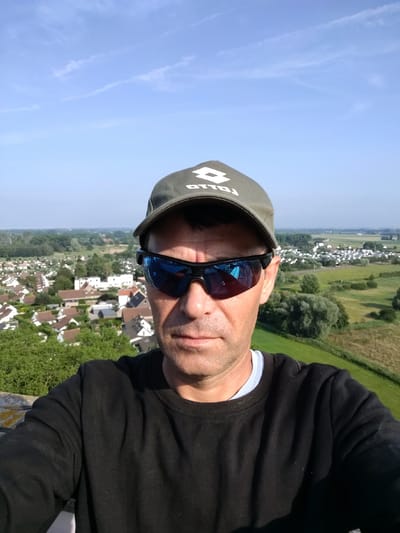 A selfie of a man wearing a dark green baseball cap with a white logo and dark sunglasses. He is looking directly at the camera with a neutral expression. The man is wearing a black crewneck sweatshirt. The background shows a landscape with a town or village, green fields, and trees under a clear blue sky with some wispy clouds. The sun appears to be shining brightly, suggesting it is daytime. The man is in the foreground, and the landscape recedes into the distance. The logo on his cap appears to be "LOTTO". The image was taken in Koksijde, Belgium.
