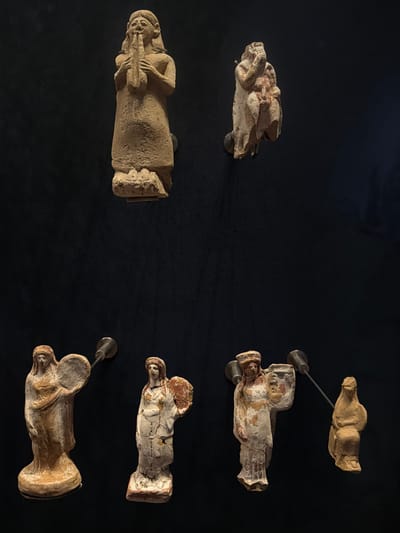 Six ancient clay figurines are displayed against a black backdrop. Two figurines are positioned at the top of the frame. The left figurine, a standing male figure with a long beard, holds a musical instrument resembling a lyre to his mouth. The right figurine is a seated or crouched figure with its face obscured.
Below these, four more figurines are arranged horizontally. From left to right, the first is a standing female figure holding a circular object, possibly a shield or tambourine. The second is a standing female figure with painted details, also holding a circular object. The third is a standing female figure holding a vessel. The fourth is a seated female figure.
The figurines are mounted on the wall with small metal rods. The lighting appears to be directed, creating shadows that enhance the three-dimensionality of the objects. The overall presentation suggests an exhibition or museum display. This scene is located at the Istanbul Archaeological Museums in Istanbul, Turkey.