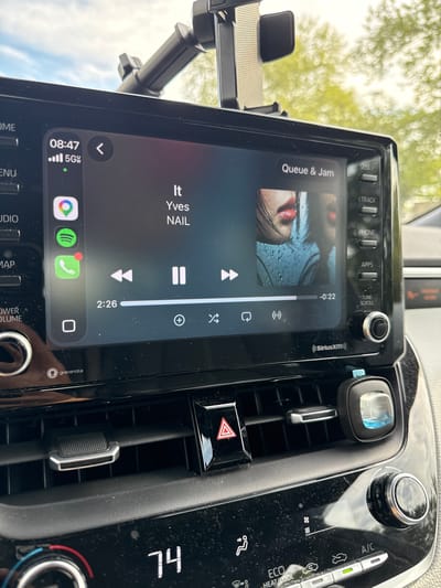 The image captures the interior of a car, specifically focusing on the dashboard and the infotainment system. The scene is set in Glen Burnie, United States of America, during daytime, indicated by the bright sky and trees visible through the windshield.
The central subject is a vertically oriented car infotainment screen displaying a music player interface. The song "Queue & Jam" by "Yves NAIL" is currently playing, with its album art showing a close-up of a person's face, specifically their lips. The progress bar indicates the song is 0:22 seconds in, with 2:26 remaining. Various music controls such as play/pause, skip, and shuffle are visible. The screen also shows the current time as 08:47 AM, a 5G network indicator, and app icons for Google Maps, Spotify, and a phone application. Along the top of the screen are touch buttons for "SEEK", "TRACK", "PHONE", "APPS", and "TUNE SCROLL". Logos for "Siriusxm" and "gracenote" are also present.
To the left of the screen, a black phone mount is attached. To the right, an air vent holds a small, light-blue air freshener or diffuser. Below the vent, a red triangular hazard light button is visible. Further right are climate control dials and buttons, with "A/C", "ECO", and "HEAT/COOL" labels, and the temperature set to "74". The dashboard and screen show some specks of dust.