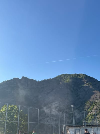 The image depicts a tall, rocky mountain in Bukovo, Bulgaria, partially covered with greenery and facing a clear, blue sky with a contrail. A metal structure resembling a fence or viewing platform is visible in the foreground, with glimpses of people standing on it. Mist or spray partially obscures the base of the mountain.