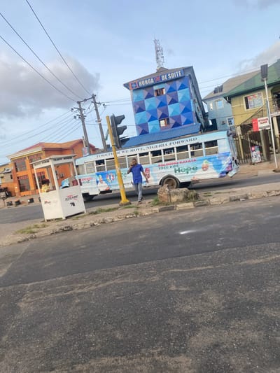 The image depicts a street scene in Agege, Nigeria. A bus advertising "Oluomo Kafata Free Ride, Renewed Agenda 2027" is parked near a traffic officer's booth labeled "Mikano". A man in a blue shirt is standing next to the bus. A building with "Hungam de Suite" signage is visible in the background, along with other buildings. Power lines and a traffic light structure are also present. The street is paved and shows signs of wear.
