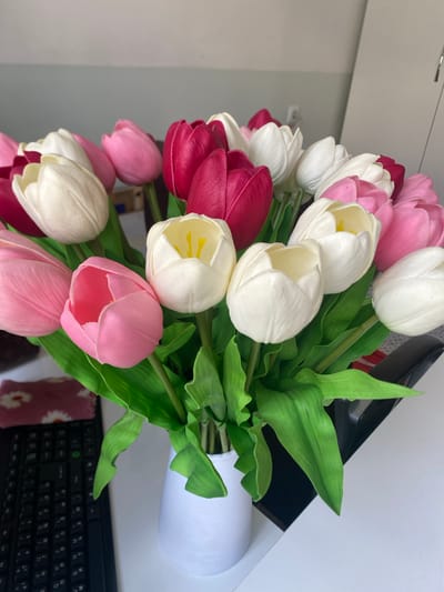 A vertical, overhead shot showcases a bouquet of artificial tulips in a white vase, set against a mirrored surface and a light-colored wall. The tulips are a mix of colors including white, pink, and a deep magenta. The arrangement is lush, with abundant green leaves.
The bouquet is positioned on a desk, with a black keyboard visible in the lower right corner of the frame. A hint of a dark rectangular object, possibly a laptop or monitor, is visible behind the flowers, suggesting an office or home workspace. The lighting appears to be artificial, casting a bright, even illumination on the scene, without any strong shadows, indicative of an indoor setting. The overall mood is serene and decorative, with the vibrant colors of the tulips providing a focal point. There is no discernible time of day or weather information, and the location cues are limited to a desk setup. No people are visible in the frame. The visible text from the keyboard is "N B M T K P m n W U H G H Y".