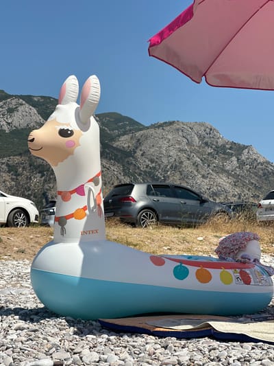 An inflatable llama float is positioned on a pebble beach in Đurovići, Montenegro, under a clear blue sky. The llama, primarily white with a light tan face and pink inner ears, features a garland of colorful balls around its neck. Its body is adorned with more colorful circular designs. A pink beach umbrella is partially visible at the top right of the frame. In the background, a line of parked cars, including a gray Volkswagen Golf, are situated on a dry, grassy area. Behind the cars, rugged, tree-covered mountains rise against the sky, indicating a coastal or mountainous region. A floral-patterned fabric is draped over the rear of the inflatable llama. A beach towel is placed beneath the float, suggesting a recreational setting.