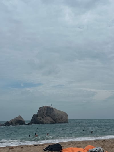 A coastal view features a cloudy sky above a sandy beach. The ocean meets the shore, with several people swimming in the water. A large, rocky formation rises from the sea near the beach, with a person standing atop one of the rocks. Orange and black items are visible on the sand, likely beach gear. The scene suggests a cloudy day at a beach with rock formations and swimming activities.