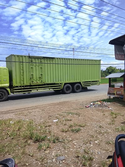 The image shows a green semi-trailer truck driving on a road, likely in Manyang Cut, Indonesia, given the context. The truck has a long, ribbed green cargo container. In the foreground, there is a roadside stall with a sign displaying "SIMPATI" and other text in Indonesian. Piles of trash are scattered on the ground near the stall. The background features a blue sky with scattered clouds, power lines, and some greenery, possibly agricultural fields and trees. The scene appears to be a typical roadside view in a rural or semi-urban Indonesian setting.