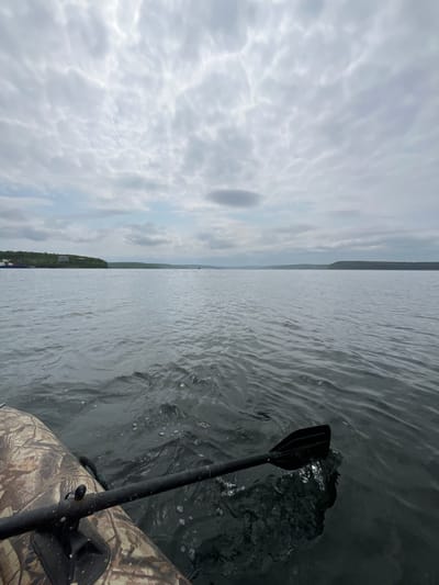 The image shows a first-person view from a camouflage-patterned inflatable boat on a calm body of water, likely a bay or inlet. A black oar is partially submerged in the water on the right side of the frame, creating ripples and small splashes. The water appears dark and slightly choppy. In the distance, a tree-covered shoreline with some buildings is visible on the left. The sky is overcast with dramatic, textured clouds. The scene is set in Sovetskaya Gavan', Russia, a port city known for its maritime activities.