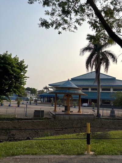 This image captures an outdoor scene in Bencongan Indah, Indonesia, likely a public space or the grounds of an institution. In the foreground, a patch of green grass is bordered by a paved walkway. A yellow and black striped pole stands prominently on the grass. Behind the grass, a low stone wall separates it from a fenced area. This fenced area contains a gravel-like ground surface with several potted plants arranged in a row.

In the middle ground, a small, ornate wooden gazebo with a blue, multi-tiered roof stands on a slightly raised platform. Behind the gazebo, a large building with a distinctive blue roof and white facade is visible. The building appears to be a modern institutional structure, possibly a school, community center, or office complex. A tall palm tree is situated to the right of the gazebo, partially obscuring the building. Several cars are parked in the lot in front of the building.

The background consists of a hazy, pale sky, suggesting it might be late afternoon or early morning. The scene is framed by the branches of trees in the upper left and right corners, with one large tree trunk visible on the right. The overall impression is one of a well-maintained, tranquil outdoor environment. There are no people visible in the image.