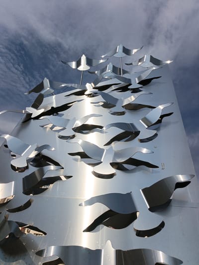 This media file features a large, metallic sculpture of birds in flight, mounted on a flat, reflective surface that appears to be part of a building or monument. The birds are abstractly rendered, with cut-out shapes that create shadows and depth. They are arranged in a dynamic composition, suggesting movement and a flock in motion. The sculpture is predominantly silver or brushed aluminum, reflecting the sky and surrounding environment.
The sky above is a mix of blue and gray clouds, indicating a partly cloudy day. The lighting suggests it is daytime, possibly late morning or early afternoon, given the bright reflections on the metal. The scene is outdoors, and the reflective nature of the sculpture indicates a polished surface, possibly stainless steel. There are no people visible in the image. The overall impression is one of modern public art, evoking a sense of freedom and natural beauty through stylized forms. The location context is Goryachiy Klyuch, Russia. No visible text is present in the image.