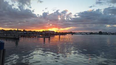 The image depicts a sunset over a harbor in Nantucket, Massachusetts, USA. The sky is filled with dramatic clouds, tinged with hues of orange, pink, and yellow as the sun sets. The water in the foreground is calm, reflecting the colors of the sky and the surrounding structures. Several wooden docks and piers extend into the water, with a few boats moored. In the background, a town skyline is visible, featuring houses and a prominent church steeple. The overall scene suggests a peaceful evening in a coastal town.