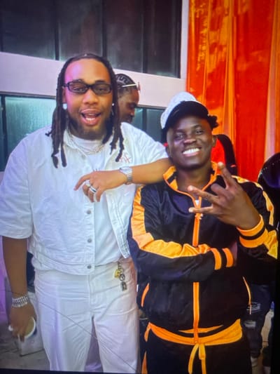 Two men smile broadly. The man on the bottom wears white pants, jacket, and sneakers. He has long dreadlocks, glasses, and multiple rings and bracelets. The other man wears a black and orange tracksuit, white hat, and silver watch. Both men are dark-skinned. They appear to be posing for a picture. The background is mostly white and orange.