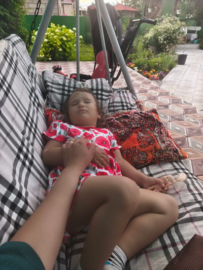 In this medium shot, a young girl is sleeping on a swing seat in what appears to be a backyard in Burayevo, Russia. She is wearing a red and white floral-patterned dress and is holding a small doll in her right hand. Her eyes are closed, and her hands are resting on her stomach. A person's hand, presumably an adult's, is also visible, gently placed on the girl's chest. The swing seat is covered with a black and white checkered cushion and a red and orange patterned blanket. In the background, there is a garden with blooming flowers, a green fence, and part of a house. A stroller is also visible next to the swing. The scene suggests a peaceful afternoon or early evening, possibly a nap time for the child. The lighting indicates it is daytime.
