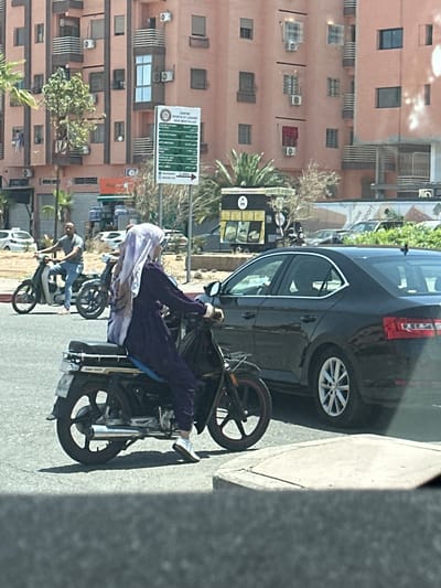 The image shows a street scene in Tassoultante, Morocco. A woman wearing a white head covering and a purple robe is seated on a moped, which is parked on the side of the road next to a dark-colored car. Another person is shown operating a similar moped nearby. The background includes buildings with air conditioning units and palm trees, as well as a street sign in Arabic and French.
