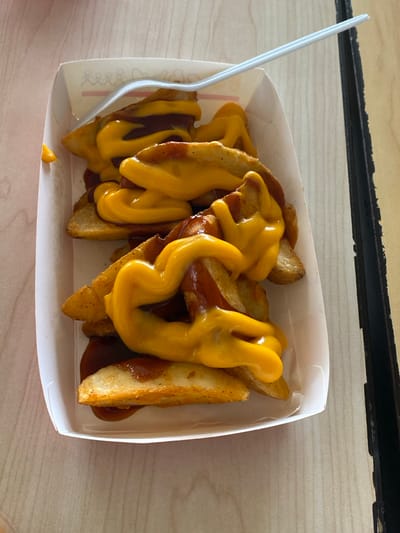 A close-up, high-angle shot displays a serving of potato wedges generously covered in two distinct sauces, presented in a white paper tray. The wedges are golden-brown and appear freshly fried, with some showing bits of potato skin. They are heavily drizzled with a dark reddish-brown sauce, possibly barbecue or chili, and a thick, bright yellow, creamy cheese sauce that forms swirls and drips over the wedges and the darker sauce.

A white plastic fork or spork rests on the left side of the tray, indicating the food is ready for consumption. A small drop of yellow cheese sauce has fallen onto the bottom of the container near the fork. The container itself has slightly curved sides and appears to be disposable takeout packaging. Faint, light pink or red, unreadable text or a design is visible on the inner bottom-left surface of the container.

The food is placed on a light-colored wooden surface, likely a table, with a dark, possibly metallic or plastic, horizontal strip visible along the top edge of the frame. No people are visible in the image, and there are no specific cues to determine the time of day, weather, or emotions. While the location context of Bekasi Regency, Indonesia, was provided, there are no visual elements within the image to specifically confirm this location.