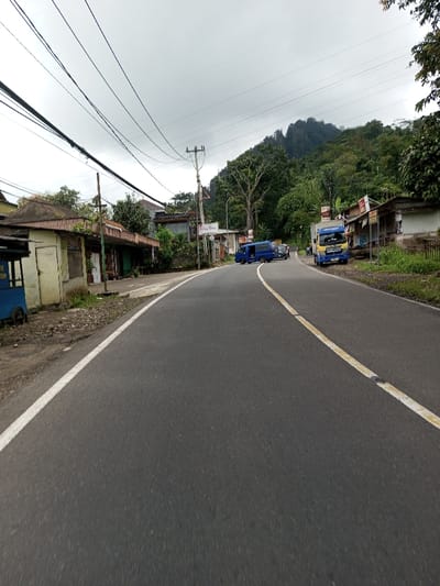 This image depicts a two-lane asphalt road curving gently to the right in Serpang Shalli, Bhutan, under an overcast sky. The road features a solid white line marking its left edge and a solid yellow line near its center. On the left side of the road, a dirt shoulder with sparse vegetation abuts a series of single-story, utilitarian buildings or storefronts, one of which is pale yellow with a dark roof and a closed door. A blue vendor cart with "KUTANG" partially visible is situated to the far left. Overhead, numerous black utility lines traverse the scene, supported by wooden and metal poles.
On the right side of the road, as it curves, a blue passenger van is parked facing away from the viewer, with other vehicles partially visible behind it. Further along, a light blue and yellow delivery truck, marked with "SOLAR," is parked facing towards the viewer. Adjacent to the road on this side are additional small structures, some with signage, and a low fence. The background is dominated by a large, densely forested hill or mountain under the uniformly gray, cloudy sky. No individuals are visible in the scene.