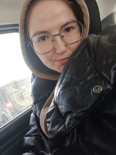 A female individual is depicted from the chest up, seated within a vehicle in Старый Шагирт, Russia. She has light skin, dark hair, and wears hexagonal-framed eyeglasses. She is dressed in a light brown hooded garment under a shiny black puffer jacket, on which a silver snap button is visible on the right side of the frame. The individual looks directly at the camera with a neutral expression, her lips slightly parted. To the left, through a vehicle window, a bright, overcast outdoor environment is visible, partially obscuring what appears to be another dark vehicle with a rectangular red and white marker in the background. The interior of the vehicle is darker, with a seat headrest visible behind the subject.