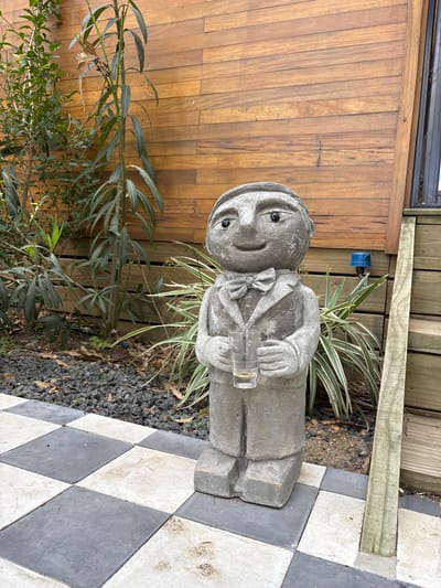 The image shows a weathered concrete statue of a man dressed in a suit and bow tie, holding a clear glass in his hands. The statue stands on a checkerboard-patterned tiled surface. Behind the statue is a wooden fence and some foliage, including a green leafy plant and a striped leafy plant. The ground around the statue and tiles is covered in small gravel and organic matter. The location is likely in a garden or outdoor space in the city of Marindia, Uruguay.