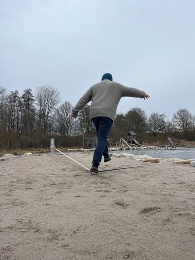An individual is captured mid-action on a slackline in an outdoor recreational area in Nohfelden, Germany. The subject, viewed from behind, wears a teal knit beanie, a grey textured sweater, dark blue pants, and dark athletic shoes, with arms extended horizontally for balance. Their left foot is positioned on a low-lying slackline, while the right foot is elevated above it, indicating a step. The slackline, composed of white and dark green sections, is anchored to a grey metal post on the left. The ground in the foreground and midground consists of light tan sand with visible footprints. In the background, a paved path, a wire fence, and numerous bare deciduous trees are visible. To the right, playground equipment including a wooden structure resembling a hut with a slide, along with various stone elements, and a dark, flat area, possibly an ice-covered pond or rink, complete the scene under an overcast sky.