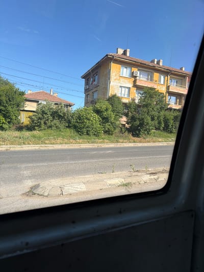 The image shows a street scene in the city of Radomir, Bulgaria, captured from inside a vehicle. The foreground is dominated by the interior of the vehicle, with a grey metal panel and a window frame visible. Through the window, a paved road with white lane markings runs horizontally across the frame. A concrete curb and a grassy verge separate the road from a sidewalk.

Beyond the verge, several buildings and trees are visible. On the right, a weathered, multi-story apartment building with a yellow facade stands out. It has a red-tiled roof, several windows, and balconies. A Bulgarian flag is hanging from one of the windows. To the left of this building, dense green trees obscure parts of another building. Further to the left, a smaller, house-like structure with a red-tiled roof and a yellow facade is partially visible behind a thick growth of trees and bushes. The sky is a clear, bright blue, with a faint contrail visible. The overall impression is of a sunny day in a residential area of the city.