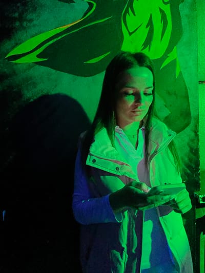 A young woman with long dark hair stands in front of a green mural in what appears to be an indoor space, possibly a club or entertainment venue. The mural depicts abstract, flowing shapes in varying shades of green, illuminated by green lighting that casts a strong glow on the scene. The woman is wearing a light-colored puffer vest over a pink collared shirt and blue sleeves. She is holding a smartphone in her hands and looking down at it, seemingly engrossed in its screen. Her expression is neutral, and her eyes are partially closed. The lighting creates stark shadows, particularly to the left of the woman, obscuring much of the background. The overall impression is of a moment captured in a vibrant, dimly lit environment. The location is identified as Kizner, Russia.