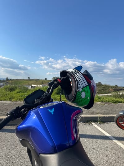 The scene depicts a metallic blue Yamaha motorcycle, identifiable by its fuel tank logo, parked outdoors in Mirandela, Portugal. A multi-colored full-face helmet with green, red, white, black, and blue comic-style graphics hangs from the motorcycle's black left handlebar. The motorcycle's digital instrument cluster and a portion of its black seat are visible. The ground consists of asphalt pavement, with a white parking line visible on the right. In the midground, a strip of green grass and weeds borders the paved area, leading to a landscape of dry grass, scattered bushes, and distant light-colored structures on a hillside. The sky is bright blue with scattered white cumulus clouds. A portion of a second motorcycle's front wheel, featuring an orange rim, is visible in the far right background.