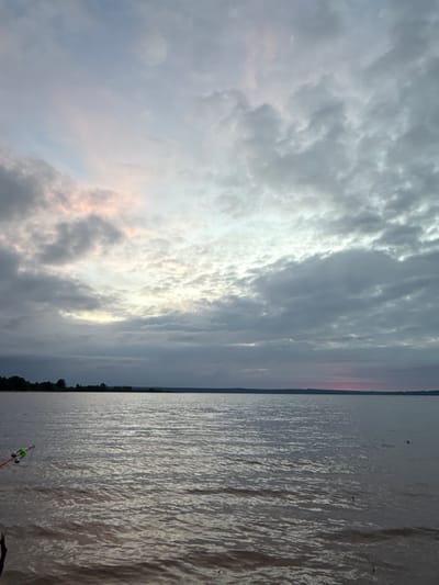 The image depicts a broad expanse of water under an overcast sky, characteristic of a natural landscape in the vicinity of Elovo, Russia. The lower two-thirds of the frame are occupied by a dark, grayish-brown body of water, showing numerous small ripples across its surface, reflecting the dull, diffused light from above. A distinct, dark landmass, likely a treeline, forms a low horizon line across the upper boundary of the water.

The upper third of the image is dominated by an extensive layer of dense, low-lying gray clouds. Subtle patches of pale pink and faint orange luminosity are visible within the clouds on the left side of the frame and a more distinct, deep pink band appears at the far right horizon, suggesting either dawn or dusk.

In the lower-left corner of the foreground, a fishing rod is partially visible, angled downwards into the water. Attached to this rod are two distinct, light-colored bell-style bite indicators, and a red section, likely part of the rod or a line component, is also present. The scene is devoid of human figures, animals, or other explicit activities, conveying a sense of stillness and solitude.
