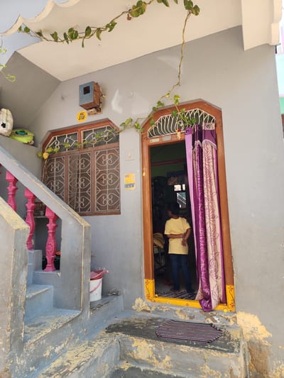The image depicts the entrance to a residence in Pedda Kammavaripalli, India. A young child, dressed in a yellow t-shirt and dark trousers, stands facing inwards within the open doorway, positioned between a patterned purple and beige curtain on the right and the interior of the home on the left. The doorway features a dark brown wooden frame with a vibrant yellow trim adorned with painted floral motifs along the bottom. A dark grey and red striped door mat lies on the worn concrete step directly outside the doorway.
To the left of the doorway, a set of concrete steps leads upwards, featuring a balustrade with prominent pink, ornate balusters and a grey handrail. The walls of the structure are painted a light grey, showing signs of wear and peeling paint, especially on the steps and lower sections. Above the steps and to the left of the door, a rectangular window with a dark brown wooden frame is fitted with white, intricate metal grilles. Above this window, a utility box, possibly an electrical meter or junction box, is mounted on the wall. A yellow sticker with "HSC 6708" is affixed below it. Further to the right of the window, near the door frame, a white light switch is visible, along with a yellow sticker displaying "103" and a QR code below it.
Overhead, a light-colored soffit or overhang is adorned with a trailing green vine with yellow-green leaves. Another section of the vine extends downwards near the window. On the upper landing, partially visible behind the balustrade, a white plastic bucket with a red handle and a green object are present. A white bucket with a pink lid is positioned under the upper part of the stairs. The overall scene suggests a lived-in residential environment with a mix of functional utility and decorative elements.