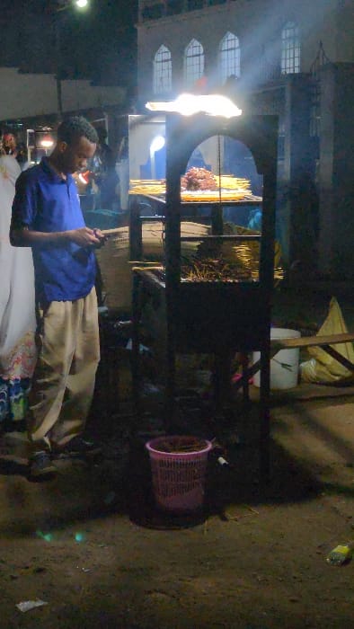 A man in a blue shirt is looking at a phone. He is in front of a glass display case with various items behind the glass. The location appears to be an outdoor market with buildings in the background. To the right, there is a pink container. The ground is dirt. The image is turned on its side.