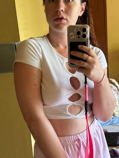 A young woman, with wet dark hair, is taking a selfie in what appears to be a bedroom. She is wearing a white ribbed crop top with cutouts and light pink sweatpants. She is holding a silver iPhone in her right hand and a pink lanyard is attached to it. A ring is visible on her left ring finger and a thin pink bracelet is on her right wrist. The background shows a yellow wall, a wooden door frame, and a bed with colorful bedding. There is no visible indication of the location being in Baan Plai Laem, Thailand.