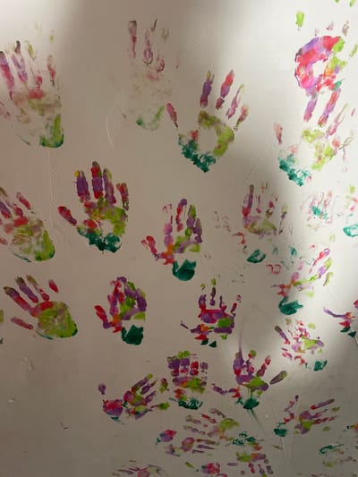 The scene depicts a light-colored, possibly white or off-white, interior wall or surface, located in the city of Kostenets, Bulgaria. Numerous colorful handprints are extensively applied across the surface. The handprints feature a blend of green, pink, purple, and red pigments, with some instances of orange and yellow. The prints vary in size and orientation, appearing to be from multiple individuals or repeated applications, with some overlapping. The surface shows minor textural variations and faint linear markings. Illumination is uneven, casting a prominent diagonal shadow across the right portion of the image. No individuals, specific objects other than the handprints, or active interactions are visible in the frame.