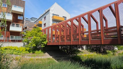 This image depicts a modern pedestrian bridge with a rusted red metal frame and mesh fencing, spanning over a small water channel in Linköping, Sweden. The bridge connects to a contemporary building complex characterized by wooden facades, white balconies with glass railings, and orange accent elements on some of the balconies. The complex appears to be residential or office buildings. Green vegetation, including tall grasses and a deciduous tree with bright green foliage, surrounds the bridge and the buildings, suggesting a well-maintained urban green space. The sky is clear and blue, indicating fair weather. There are no people visible in the image. The scene is captured from a low angle, looking up towards the bridge and buildings.