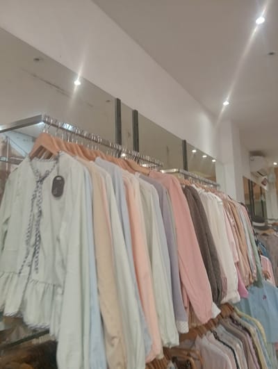 The image depicts a clothing store in Larangan, Indonesia. The scene is dominated by racks of women's clothing, mostly in pastel colors, hanging on wooden hangers. The garments appear to be blouses or dresses with various necklines and sleeve styles. The store interior is brightly lit by ceiling lights, some of which are visible in the upper portion of the image. A large mirror is also present, reflecting the store's interior and the clothing racks. In the background, an air conditioning unit is mounted on the wall. The overall impression is of a well-stocked and inviting retail space.