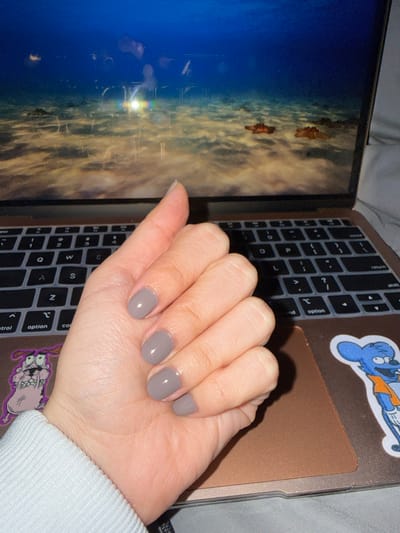 A hand with neatly painted gray nails rests on top of a laptop. The laptop features a gray keyboard cover, and stickers of cartoon characters adorn the surface. The screen displays a scene with a blue and tan color palette, potentially depicting a landscape or underwater environment. A white sleeve is visible on the right, suggesting the person wearing the hand is dressed casually.