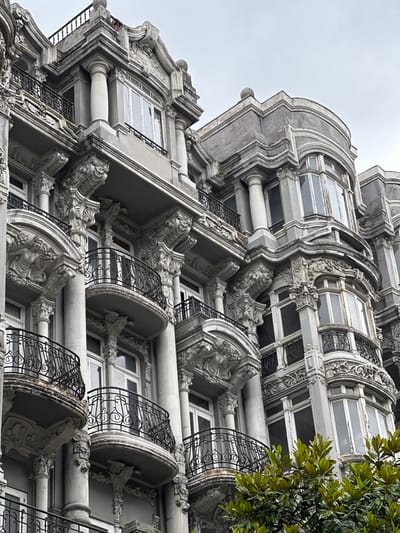 This is a close-up, low-angle, outdoor, eye-level shot of a grey, ornate, multi-story building with classical architectural elements. The building features multiple balconies with decorative black iron railings. The facade is adorned with elaborate sculptures and carvings, including figures of women and ornate corbels supporting the balconies. The windows are tall and appear to be made of wood with multiple panes. Some areas of the building show signs of weathering and wear. In the foreground, the vibrant green leaves of a tree partially obscure the lower portion of the building. The sky above is overcast and grey. The building is located in Oviedo, Spain.