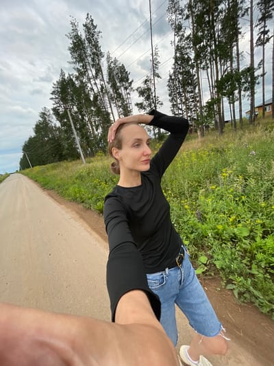 A woman with blonde hair pulled back in a bun, wearing a black long-sleeved shirt and distressed denim shorts with a black belt, stands on a dirt road in Подшивалово, Russia. She is looking to her right, with her left hand resting on the back of her head. The road stretches into the distance, lined on the right by a field of green grass and yellow wildflowers, with tall pine trees and utility poles beyond. A glimpse of a wooden house is visible through the trees in the background. The sky is overcast with clouds. The photograph is taken from a low angle, with the woman's arm and hand in the foreground.