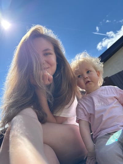 This low-angle, upward-facing image captures an adult woman and a young child against a clear blue sky in Suzdal, Russia. The photo appears to be a selfie taken outdoors during bright daytime, with a prominent sun flare visible in the lower left corner.
In the foreground, an adult woman with long, light brown hair is looking towards the camera with a gentle smile. Her hair frames her face, and a hand is visible near her chin. Above her, a young child with fair skin, curly blonde hair, and striking blue eyes looks directly into the camera with a curious and slightly smiling expression. The child is wearing a light pink long-sleeved shirt on which the word "Happy" is embroidered in white script on the chest. The child's body is positioned above the adult's, suggesting they are being held or are leaning over the adult.
The background features a vast, bright blue sky with a few scattered white clouds and a distinct white contrail from an airplane. A dark structure, possibly the roofline of a building, is partially visible in the upper right. The overall mood conveyed is one of warmth, happiness, and a close, affectionate interaction between the two individuals under a sunny sky.