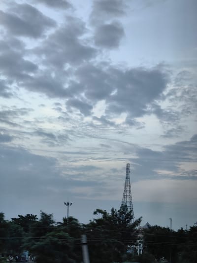 The image shows a wide shot of a landscape in Baldhi, India, with a prominent cellular tower in the center. The sky is overcast with scattered clouds, casting a muted light over the scene. In the foreground, a dense line of dark green trees runs horizontally across the frame, partially obscuring the view of what lies behind. To the left of the cellular tower, a tall lamppost with multiple lights is visible amongst the trees. Further in the distance, a few other structures and what appear to be more cellular towers can be seen. The bottom portion of the image shows a blurry area that suggests movement, possibly from a vehicle. The overall impression is one of a somewhat hazy, late afternoon or early morning light.