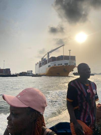 A cargo ship named “Triada Vistoria” is visible on the water near a port. People are on a boat, looking towards the vessel. The sky is overcast, with a bright sun shining through. One person wears a pink hat, while another has a black, red, and white striped shirt. The water is choppy, indicating movement. Shipping containers are stacked in the port area.