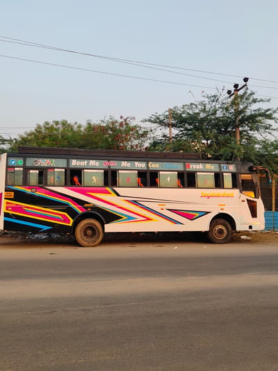A white passenger bus, designated "Jayalakshmi" and displaying license plate AP03 U.9485, is parked stationary on the side of a paved road in Anantapur, India. The bus exterior is white with prominent geometric designs in black, pink, blue, yellow, and orange across its lower and mid-sections. Above the windows, the text "Beat Me Hate Me You Can Never Break Me" is visible in various fonts and colors. Social media icons (Twitter, WhatsApp, Facebook, Instagram) and a "DJ Naipa Music" sticker are present near the front upper section. The windows feature silhouettes of individuals engaged in various activities, including dancing and sports. The bus tires appear worn and dusty. No individuals are visible on or around the bus.

The environment includes a dusty asphalt road in the foreground and beneath the bus. Behind the bus, dense green trees and shrubs are visible, some exhibiting reddish foliage. Utility poles with overhead power lines traverse the upper portion of the scene against a clear, light blue sky. On the far right, partially obscured by the bus's rear, a structure made of blue corrugated material is present. Debris, including discarded paper, is scattered on the ground adjacent to the bus.