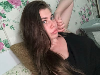 The image shows a woman with long, dark brown hair lying on her side, propped up by a patterned pillow. She is wearing a black top. Her right arm is bent, and her hand is resting on her cheek. She is looking towards the camera with a neutral expression. The background consists of floral wallpaper with pink roses and green leaves. A crib is partially visible on the right side of the image. There is no indication of the location being in the city of Мещёры, Russia.