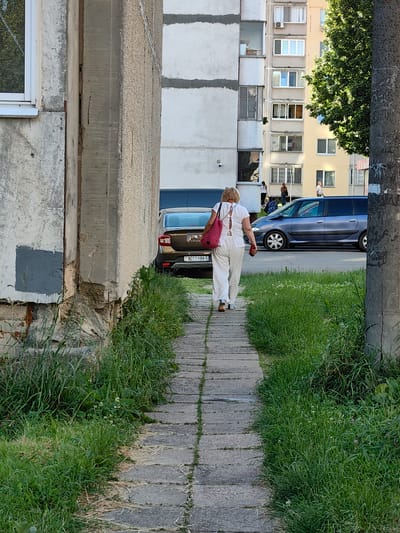 A woman walks down a paved pathway lined with overgrown grass. She is wearing a white top and white pants and carrying a red bag over her shoulder. Behind her, a brown car is parked. Further in the background, a blue minivan is driving away. Several buildings are visible, including a multi-story apartment building with many windows. The scene appears to be outdoors, possibly in a residential area. The time of day seems to be daytime, with bright sunlight casting shadows. There is no discernible weather information. The license plate of the brown car has the text "9011 HM-5".
