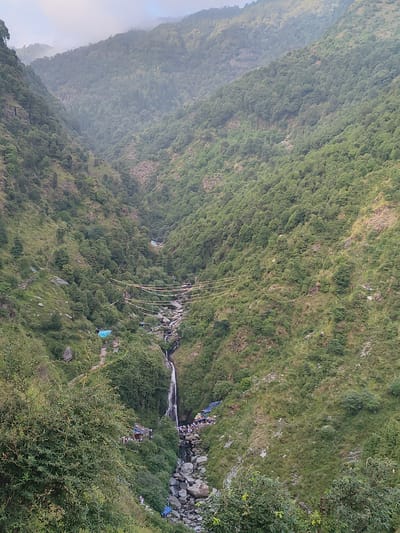 This image captures a view of Bhagsu Waterfall, a popular tourist destination in the city of Bhagsu, India. The waterfall cascades down a rocky slope amidst lush green mountains. In the foreground, dense vegetation lines a pathway leading towards the waterfall. Further down, a stream flows through a rocky bed, with several small blue tents and makeshift shelters dotting the landscape. A gathering of people is visible near the base of the waterfall, suggesting a busy tourist spot. Decorative strings of colorful flags are strung across the gorge, adding a festive touch to the natural scenery. The surrounding mountains are covered in dense green trees and vegetation, with some areas appearing rocky and rugged. The sky is overcast, contributing to a diffused light.