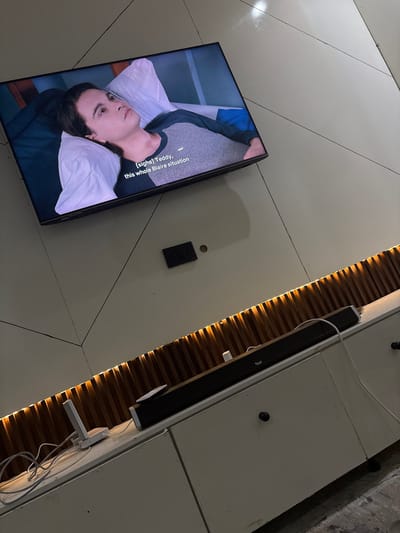 The image shows an indoor scene with a television mounted on a wall in Iddo Tudunwada, Nigeria. The television displays a scene of a person in bed. A sound bar is mounted on the wall beside the television, with a decorative panel behind it. The wall is composed of several large, flat tiles. The lighting appears to be indirect.