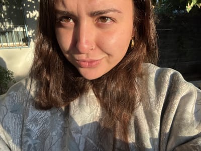 A woman with light brown hair and fair skin looks directly at the camera, which is positioned above her. She has brown eyes, a nose piercing, and is wearing a patterned gray top. Behind her, sunlight casts shadows on a white wall and a window with bars. Some green foliage is visible on the left side of the frame. The image is rotated counter-clockwise.