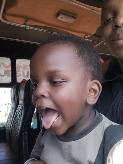 A close-up captures a young child with dark skin, cropped short hair, and their mouth wide open, tongue extended in a playful expression. Seated inside a vehicle with cushioned, stitched seating, the child is positioned next to an adult whose face is partially visible at the top right of the frame. The interior of the vehicle, including a window displaying the word "Jesus", is visible. The lighting suggests daylight.